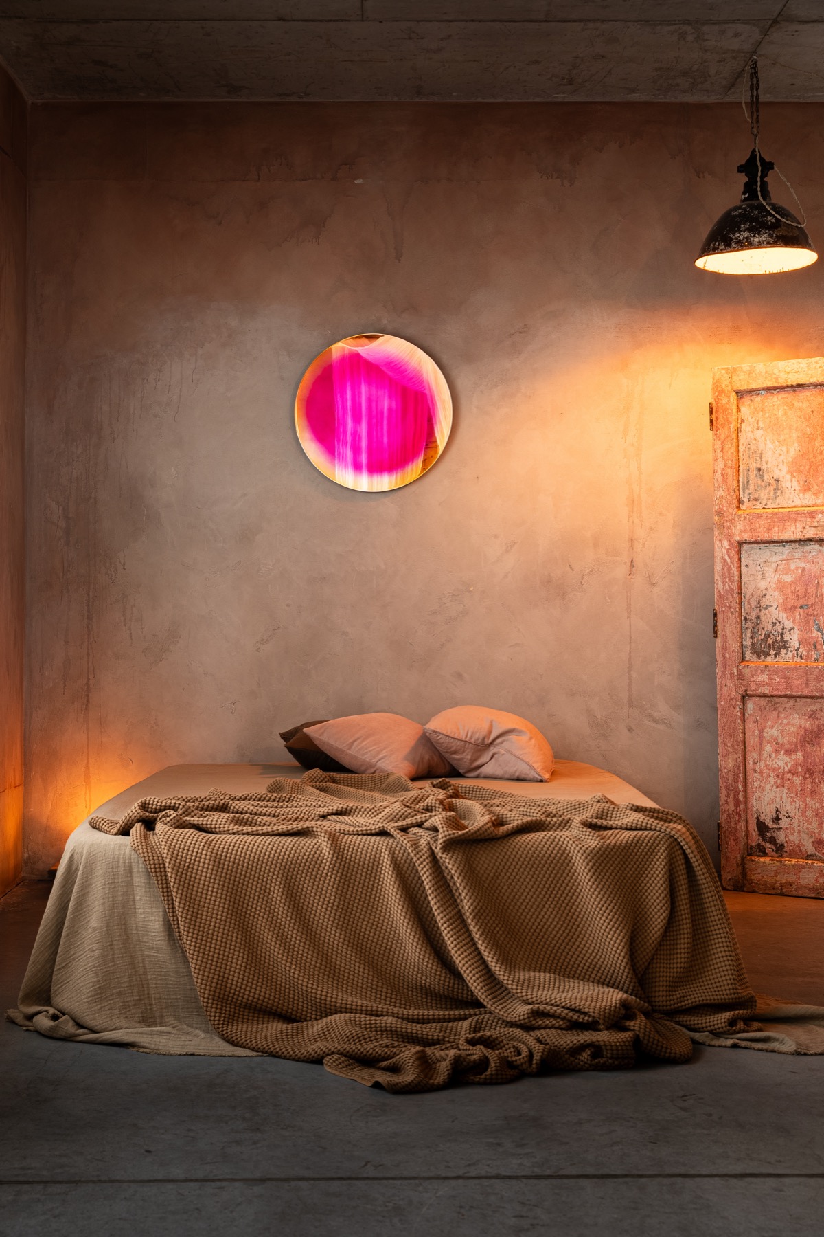The Saturn mirror with a vivid pink ring framing an amber center, hung above a bed in a warm plastered room