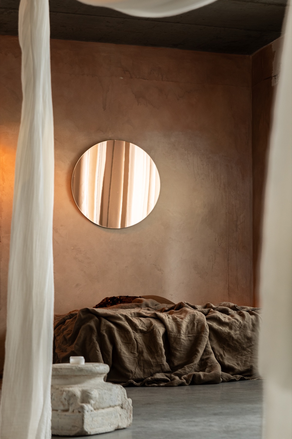 A round mirror lacquered in solid copper, reflecting curtains in an earthy plastered room